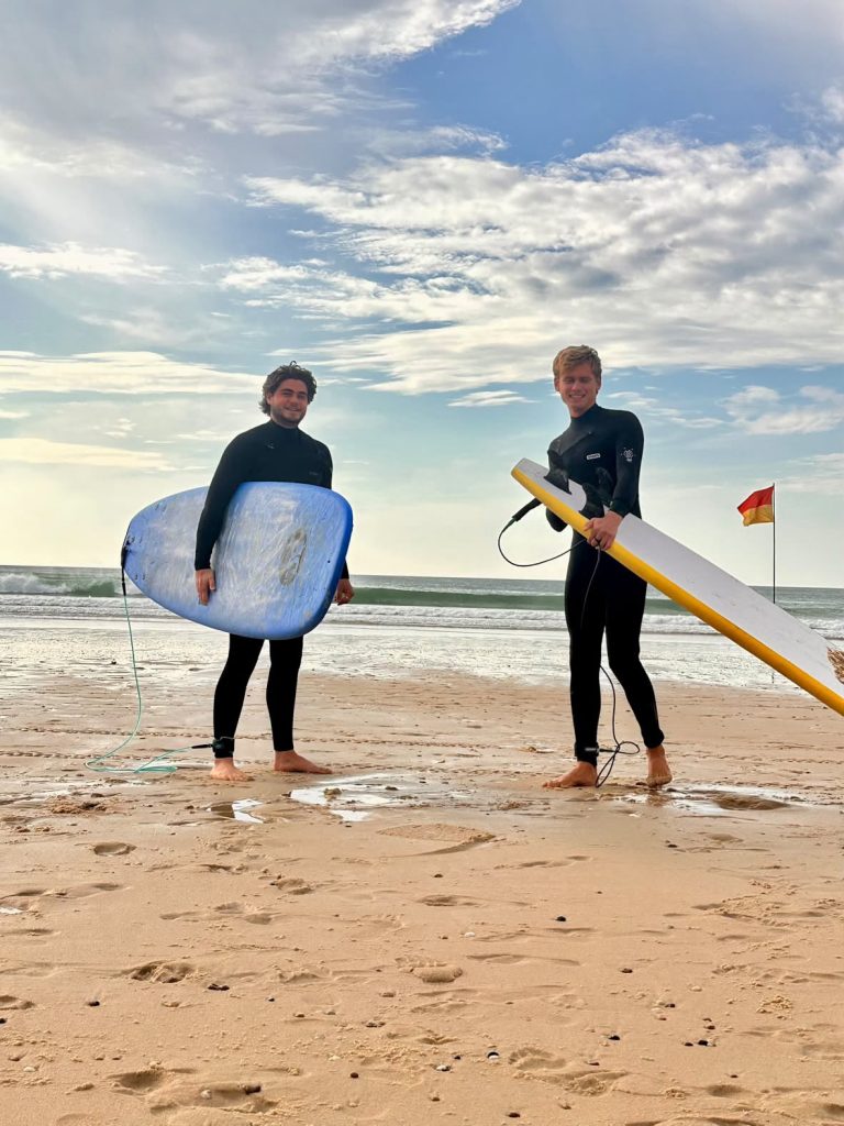 Deux surfeurs souriants en combinaison, planches à la main, au bord de l’eau